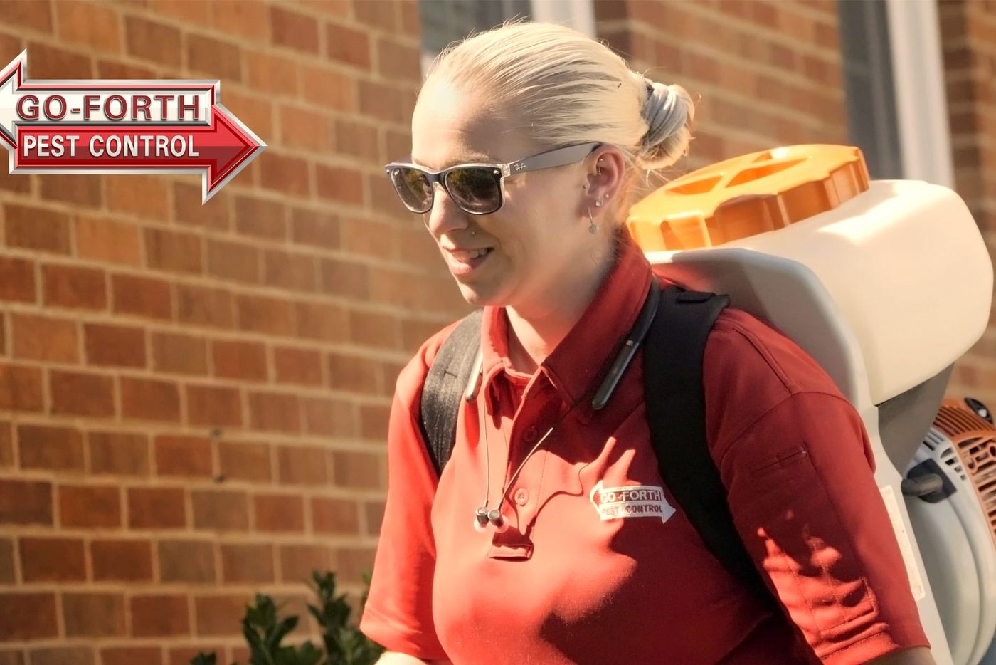 A Go-Forth technician in sunglasses carrying a backpack sprayer along a brick home
